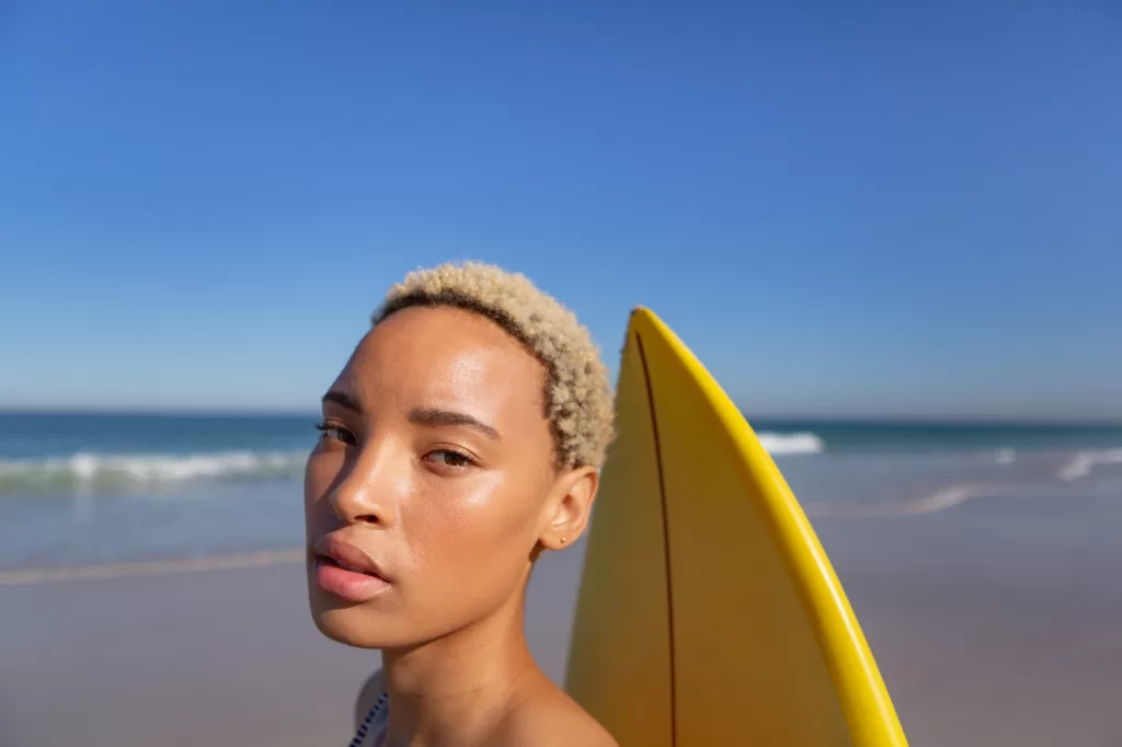 Close-up of beautiful African american woman standing with surfboard on beach in the sunshine